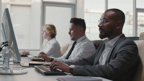 Medium slow motion shot of three multi-ethnic soft developers in formalwear spending workday in office working on desktop computers