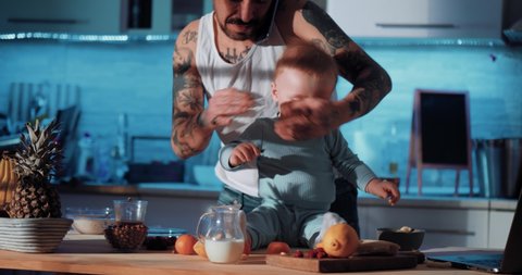 Tattooed dad wipes the baby's hands and talks on the phone. Dad and baby in the kitchen, preparing food and using laptops. Baby on the table	
