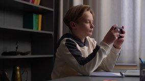 Close-up of handsome Caucasian pupil boy playing video game on mobile phone sitting at desk with studying book, workbook, laptop near window during rest, side view. Tracking shot in slow motion.