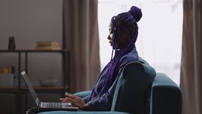 black woman is communicating by video call on laptop, sitting at home at weekend, sitting on couch in living room and talking, medium female portrait in apartment, side view against window
