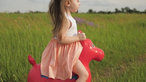 Happy little girl is playing in park and jumping on an inflatable toy horse. Kid plays on playground with his favorite toy horse. Little girl, daughter playing in field outdoors. Family and childhood.
