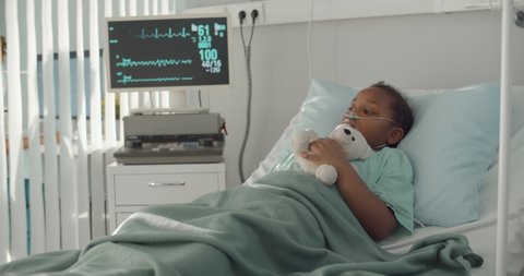 Afro-american kid lying in hospital bed with oxygen tube and plush toy. Portrait of sick and sad african child patient resting in bed at hospital ward with modern equipment