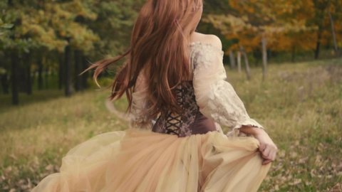 Happy joyful beautiful young woman in a medieval dress is runing in forest, enjoying nature. Slow motion. brown beige vintage dress. Long red hair fly flutter in wind. Cheerful girl back rear view.