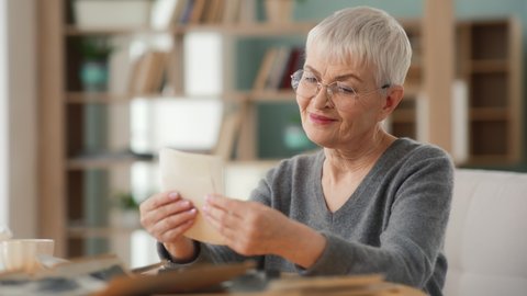 Senior woman in glasses looking at old photo album closeup. Mature female holding nostalgic memory at comfort modern flat. Casual elderly life and wrinkled skin of hands. Biography or history concept