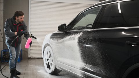 Male car expert covering dark luxury auto with foam at washing service. Mechanic doing all preparation for proper cleaning of modern vehicle.
