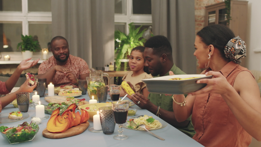 Medium PAN shot of big joyful afro-american family having dinner at home together, chatting and exchanging plates having good time celebrating special occasion
