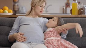 Happy, peaceful family concept.Loving little girl laughing and chatting with her pregnant mother while sitting on the sofa. Slow motion video.Close up.