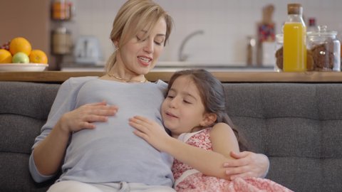 Happy, peaceful family concept.Loving little girl laughing and chatting with her pregnant mother while sitting on the sofa. Slow motion video.Close up.