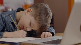 The little boy uses a laptop to make a video call with his teacher. The little boy who is studying remotely online writes the lessons the teacher told in his notebook. Distance education concept.Close
