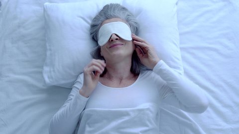 Satisfied greyhaired female enjoying nap in bed, putting on eye mask, recreation