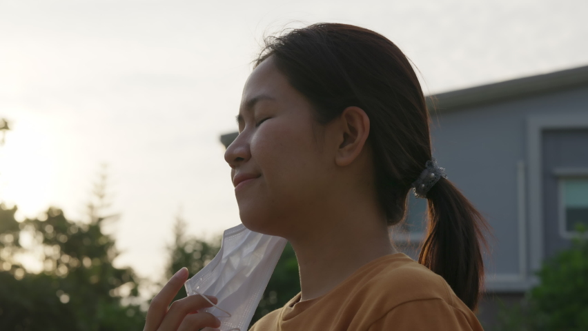 Close up Young adult asian hipster attractive woman take off medical face mask breathe free deeply and smile on sunset evening outdoor in pandamic end. Back normal life pull off mask in asia people. Royalty-Free Stock Footage #1069914028