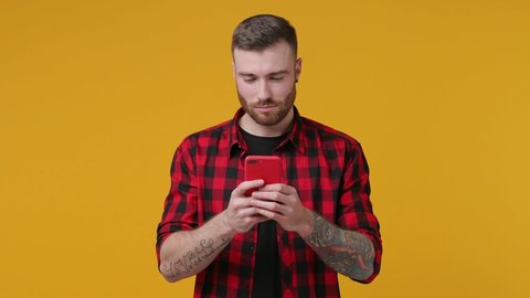 Smiling cheerful bearded tattooed young man 20s years old in red checkered shirt isolated on yellow background studio. People lifestyle concept. Tattoo translates life as a fight. Using mobile cell phone typing sms message browsing.