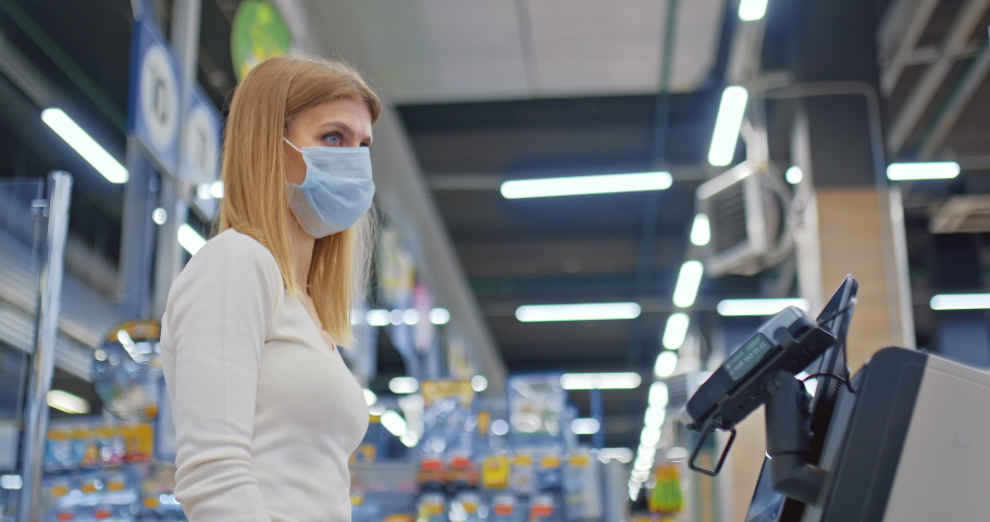 Woman in a medical mask pays for purchases in a contactless way. Woman puts a phone to a payment terminal, nfc. Self-service checkout in the supermarket. Shopping during the coronavirus period, covid | Shutterstock HD Video #1069915075