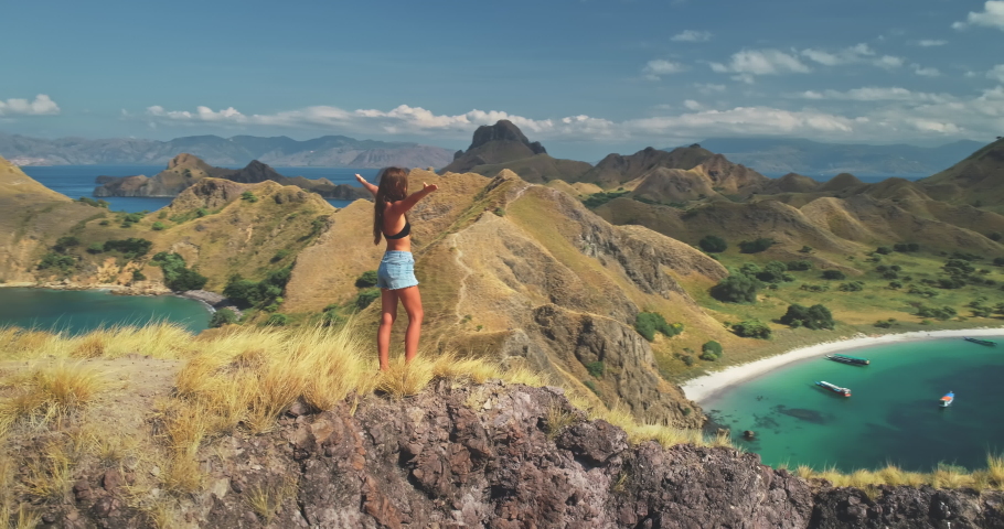 Young woman rising hands on mountain top above sea aerial. Summer nature landscape. Girl tourist standing on cliff at seaside. Lady admires tropic seascape with ships, boats. Active tourism lifestyle Royalty-Free Stock Footage #1069918390