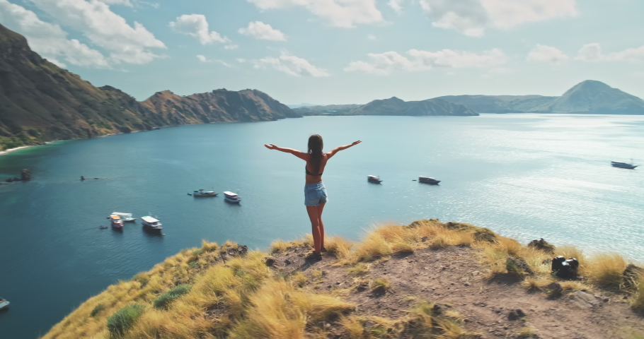 Young woman rising hands on mountain top above sea aerial. Summer nature landscape. Girl tourist standing on cliff at seaside. Lady admires tropic seascape with ships, boats. Active tourism lifestyle | Shutterstock HD Video #1069918390