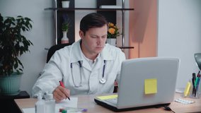 A male doctor sitting at a table writes data from a computer on a sheet of paper.