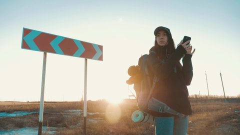 A young woman traveler uses her phone to choose the right road. Hiker woman with backpack choose in a road fork between two different path directions at the sunset. Concept of choose the correct way.