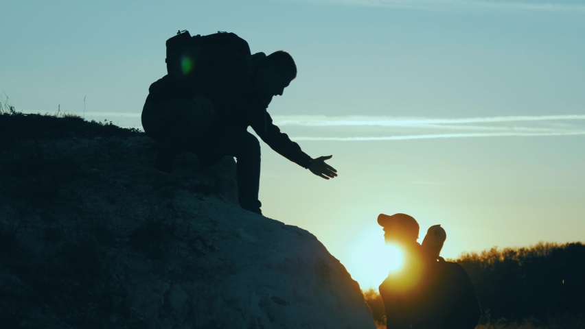 Helping Hand. Man giving hand a teammate to help her to climb the mountain. Help and assistance, hand reaching out to help friend at beautiful sunset sky. Hiking and team work concept. People helping.
