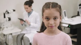Medium closeup PAN portrait with slowmo of pretty little girl smiling to camera standing at ophthalmologist office while female doctor sitting at workplace using digital tablet in background - Powered by Shutterstock - Get 15% off with code: PIKWIZARD15