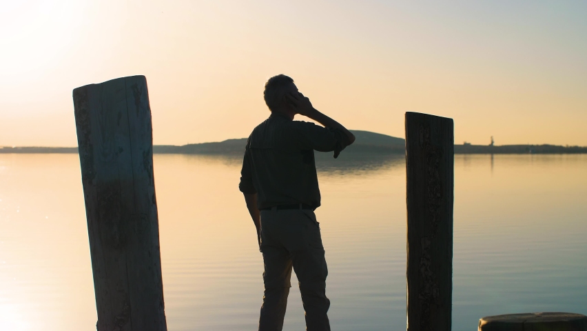 Frustrated and angry man talking on smartphone and throwing it in the lake. Silhouette of an annoyed businessman standing on a pier and throwing his phone in the water. Slow-motion, 4K. Royalty-Free Stock Footage #1069928611