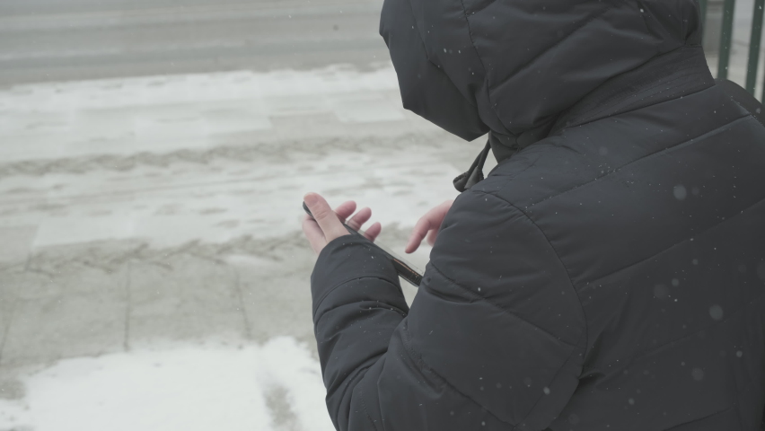 Snow is falling, cold. A girl in a warm black parka is standing next to the road and is trying to call a taxi using an app on her phone