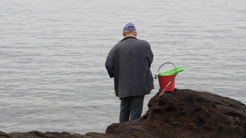 Back view of elderly fisherman fishing outdoors on sea during daytime. Bucket, and fishing rod.