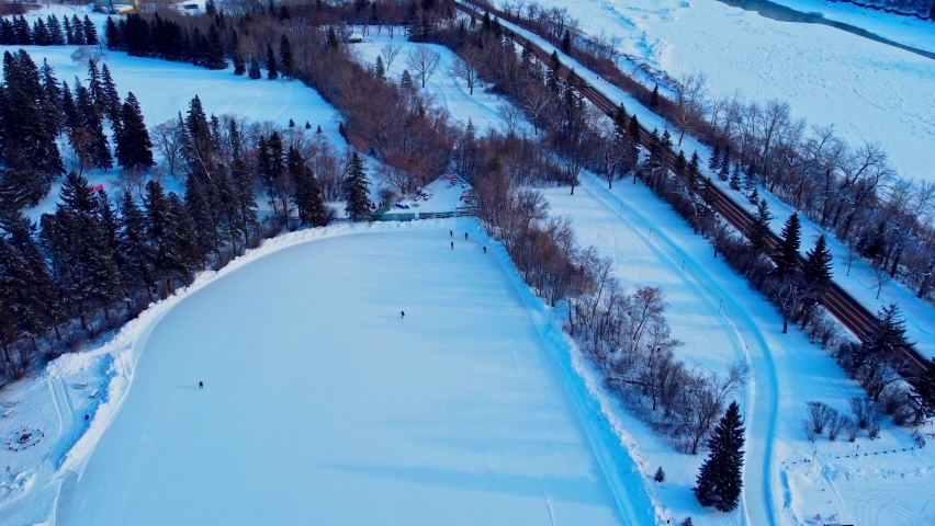 4k Winter aerial flyover drop sunset reflects shadow trees snow covered skate path next to roadside access parked cars connected to largest outdoor manmade ice rink adults skating EDM Victoria Park5-6
