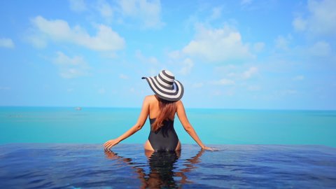 Back of slim female in swimsuit and summer hat enjoying in stunning view on sea skyline from infinity swimming pool, luxury holiday concept, full frame