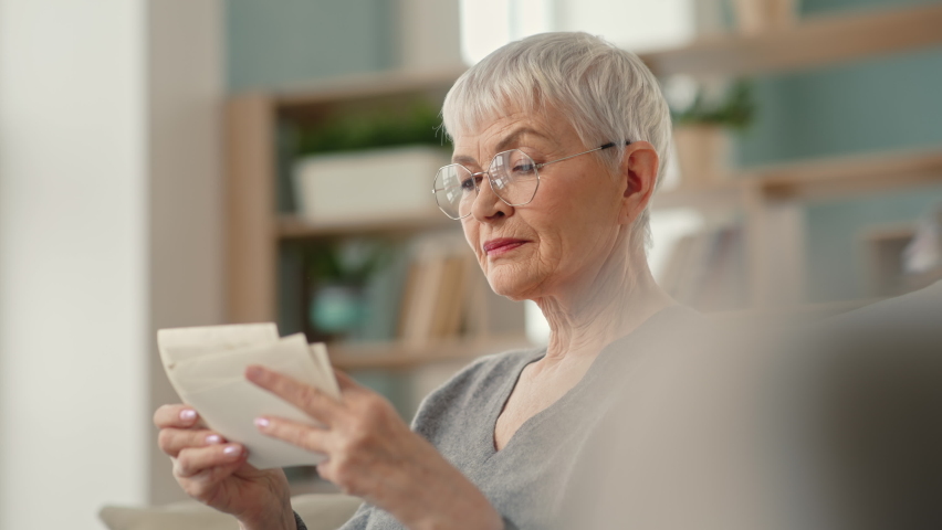 Senior 70s woman in glasses looking at old photo album close-up. Adult female holding nostalgic memory at cozy modern home. Casual elderly life and wrinkled skin of hands. Biography or history concept Royalty-Free Stock Footage #1069941208
