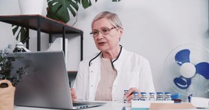 Senior Caucasian pharmacy doctor woman in lab coat showing vaccine flask to remote client online via laptop webcam call.