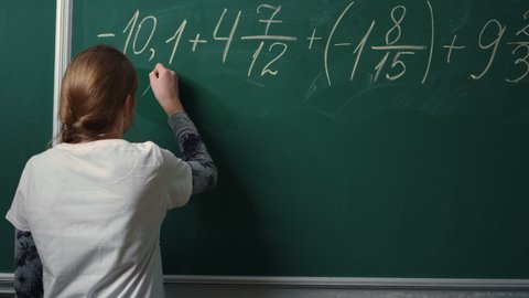 Back view of school girl doing math task on blackboard in class. Young student writing complex mathematical formula. Unrecognizable teenage girl solving example indoors.