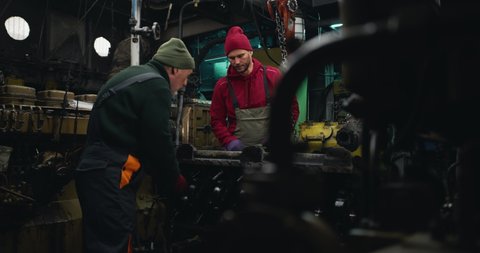 Zoom in view of adult male mechanic assisting senior man while fixing motor in engine room of ship