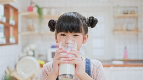 Portrait of Asian little cute kid holding a cup of milk and drinking in kitchen at home. Young preschool girl child or daughter messing up milky on moustache and lips then looking at camera in house.