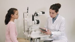 Medium shot of little girl answering female ophthalmologist questions having doctor appointment in modern clinic. Woman making notes on digital tablet during consultation - Powered by Shutterstock - Get 15% off with code: PIKWIZARD15