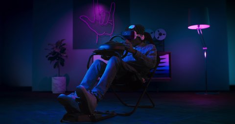 A guy is playing racing game in VR. He is sitting on the gaming chair with a steering wheel and pedals. A guy wears VR headset. Chair is located in the living room with red and blue neon lighting. 
