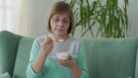 Elderly woman eats yogurt sitting on the couch at home