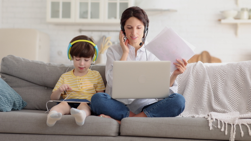 Freelancer mother sitting on couch at home office during lockdown, remote work on laptop. Child using tablet, playing in game at tablet, sitting with mom on sofa. Family, telework, e-learning concept. | Shutterstock HD Video #1069955902