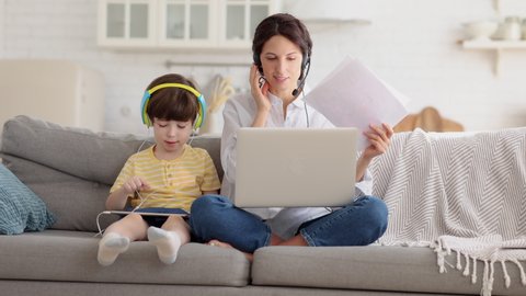 Freelancer mother sitting on couch at home office during lockdown, remote work on laptop. Child using tablet, playing in game at tablet, sitting with mom on sofa. Family, telework, e-learning concept.