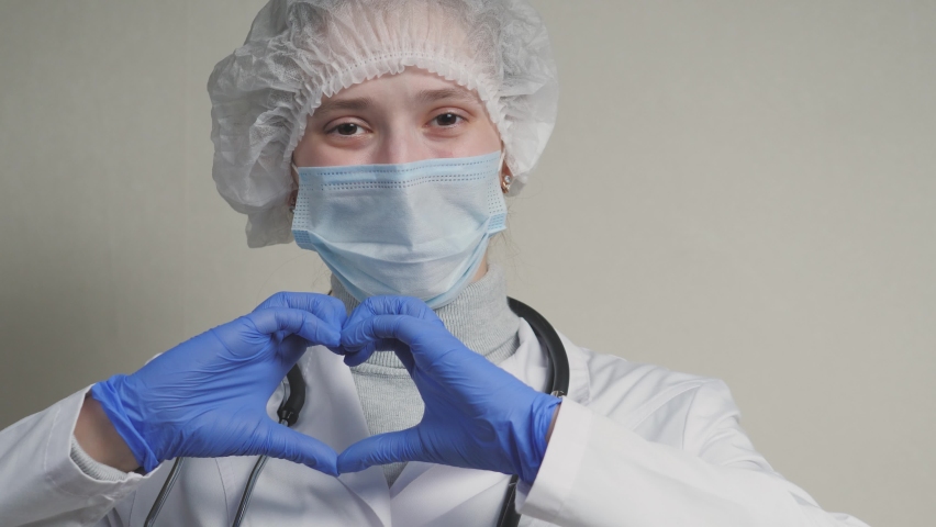 A doctor in a blue mask and gloves smiles and shows his heart, a professional cardiologist in a health clinic, an appointment with a pediatrician in the office, a good nurse, coronavirus pandemic Royalty-Free Stock Footage #1069963216