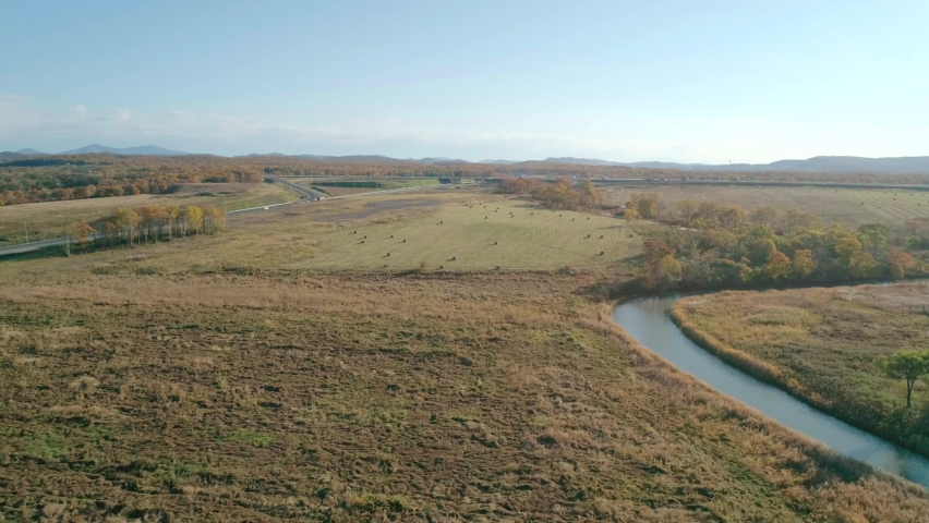 Aerial landscape of a rural nature. River, green trees, blue sky, hay bales field, roadway with traffic on the background. Day time. Camera moves right.