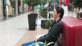 A young impatient woman is sitting on a sofa and holding a protective mask at her hands, in the waiting room, looking around. Side view. Real time. The concept of waiting.