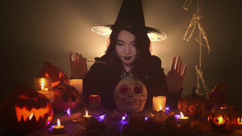 Young witch in hat and dark mantle is doing magic over skull among candles and pumpkins on black background. Pretty brunette is conjuring with joss stick surrounded by smoke and Halloween decorations.