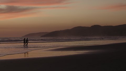 Slow motion of silhouette of distant couple walking on ocean beach at sunset , San Blas, La Libertad, El Salvador