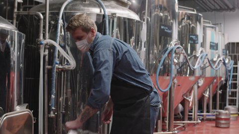 Slow-motion medium track shot of strong brutal man in protective mask working at brewery carrying heavy beer barrel