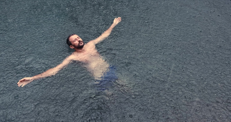 Young man floating on the back in the dark water of simming pool, ocean or lake under the rain drops