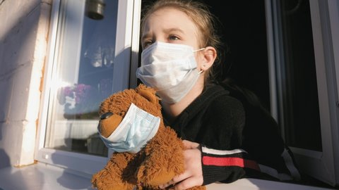 Quarantine. the threat of coronavirus. Sad child and his teddy bear. Bored girl wears a medical mask in home quarantine coronavirus looks out of the window. Epidemic covid 19 prevention concept.
