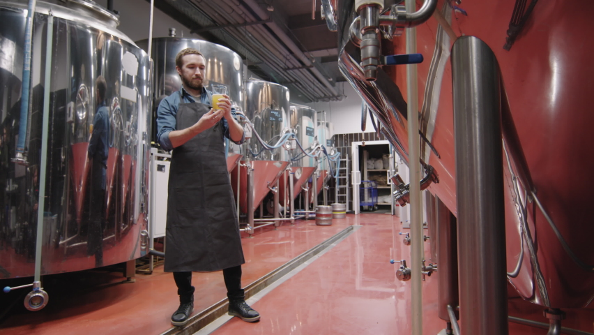 Slow-motion full PAN shot of professional brewery worker holding glass of fresh beer just produced at factory looking at it checking quality | Shutterstock HD Video #1069976035