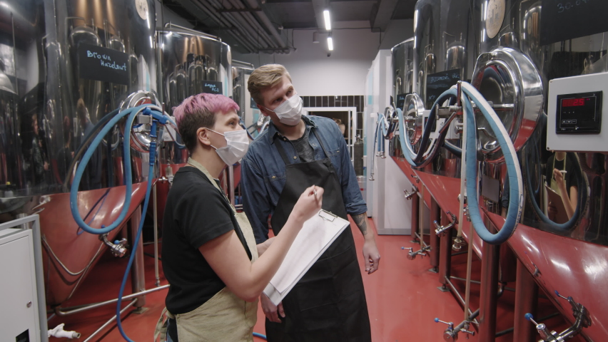 Slow-motion medium shot of brewery experts controlling beer production process checking equipment quality and making notes on tablet