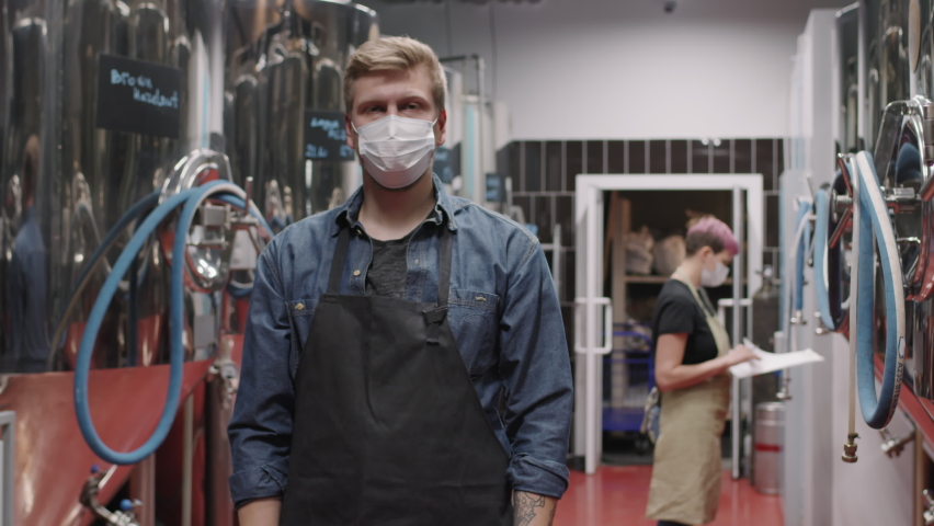 Slow-motion medium portrait of brutal brewery expert in protective face mask standing with hands folded looking at camera while female colleague controlling beer production process in background