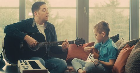 Father and son playing on guitar at home. Dad teaches child to play guitar. Music hobby, Create, learning own songs. SHARE THE LOVE, play our game. 4K video.